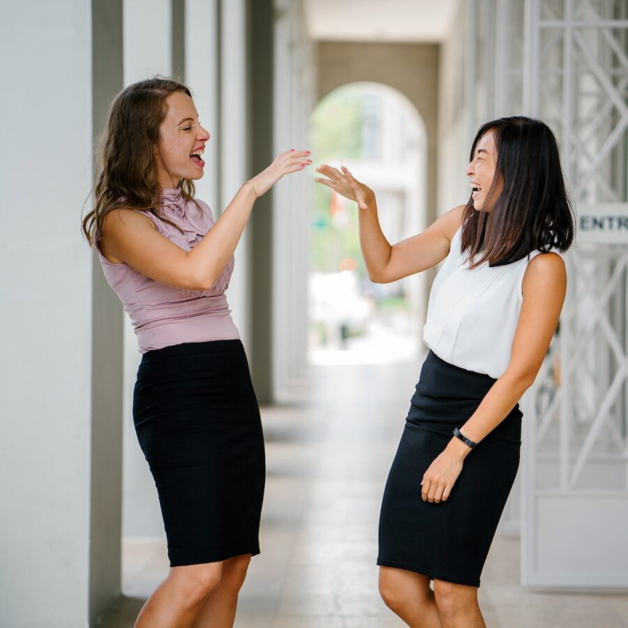 woman wearing white sleeveless top and black pencil skirt facing woman wearing pink sleeveless top and black pencil skirt leaning on wall